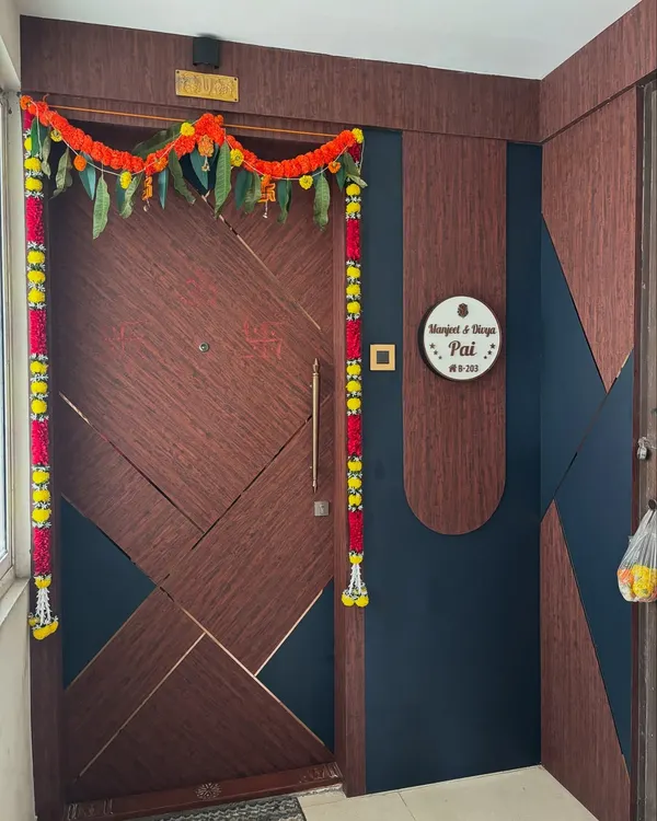A beautiful main door with a geometric pattern, combining dark wood grain and deep blue panels with fine gold inlay. The entrance is adorned for a housewarming ceremony, showcasing a proud moment for the homeowners.