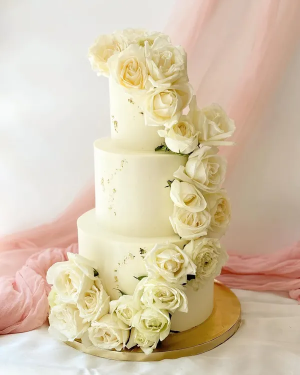 An absolutely stunning three-tier wedding cake covered in a cascading waterfall of fresh white roses. The simple, elegant design is enhanced with subtle gold leaf flecks for a touch of luxury.