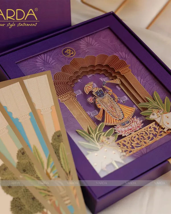 This luxurious Shreenathji-themed wedding invitation is presented in a rich purple box. The intricate 3D pop-up of Lord Shrinathji, complete with Pichwai art elements, creates a stunning and divine announcement for your wedding.