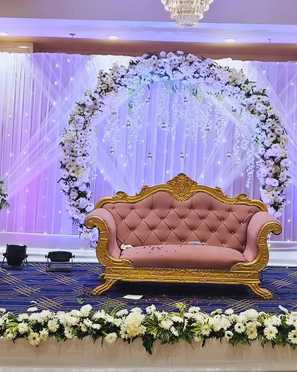 We create elegant and modern stage designs for your ring ceremony or reception. This setup features a circular floral arch with white roses and hanging crystals, a classic pink and gold sofa for the couple, and a backdrop of white drapes with fairy lights.
