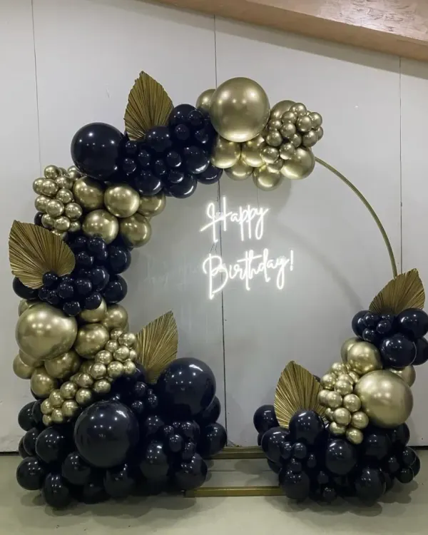 This is a high-end, modern birthday setup featuring a circular frame. The combination of matte black and chrome gold balloons, a custom neon sign, and decorative leaves creates a very sophisticated look.
