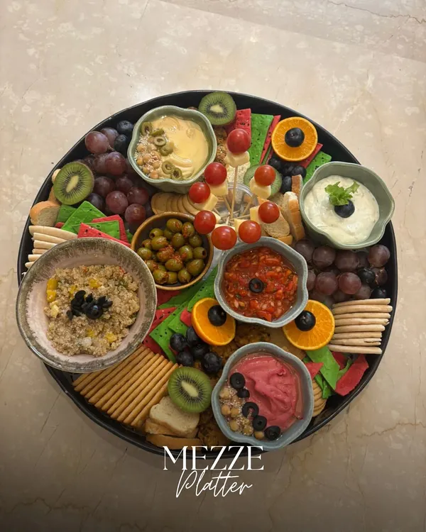 A stunning overhead view of our classic Mezze Platter. It's a colorful mosaic of beetroot hummus, classic hummus, tzatziki, olives, fresh fruits, and assorted crackers, perfect for any social gathering.