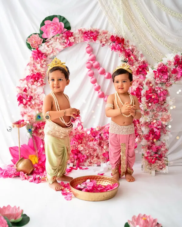 Two adorable little Krishnas. These twin boys are dressed in traditional attire, standing in front of a beautiful pink floral backdrop, making for a divine Janmashtami celebration.
