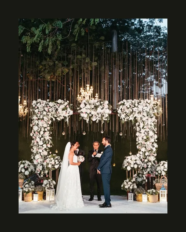 A breathtaking view of an outdoor Catholic wedding ceremony. The couple stands under a magnificent floral arch, exchanging vows in a setting that feels both grand and deeply intimate.