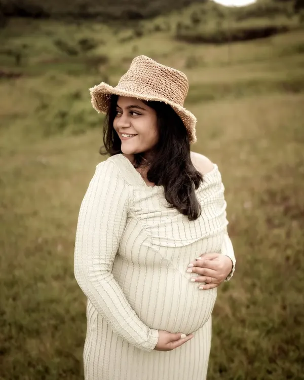 A beautiful, happy portrait of the mom-to-be. Her smile is radiant as she cradles her bump, wearing a straw hat that adds a touch of rustic charm to this outdoor solo shot.