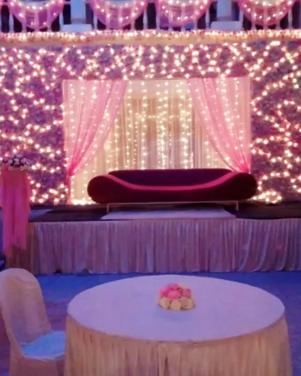 A romantic reception stage featuring a wall of pink flowers illuminated by fairy lights, with a simple red divan sofa, creating a dreamy photo backdrop.