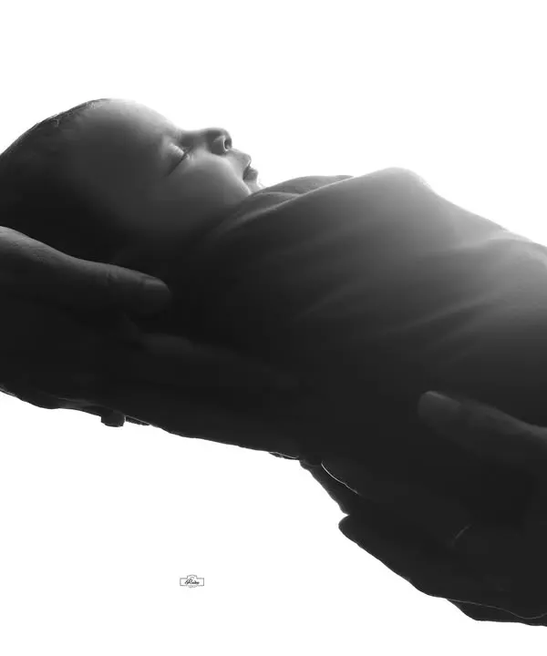 A beautiful black and white portrait showing a newborn held securely in their parents' hands. This high-contrast image emphasizes the delicate size of the baby and the protective love of the family.