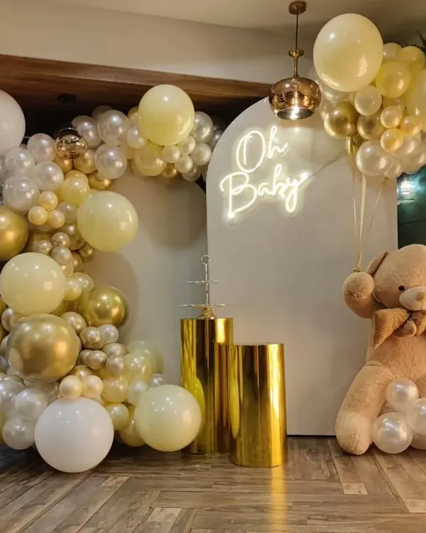 This is my popular 'Oh Baby' shower decoration. It features a beautiful balloon arch in white, cream, and chrome gold, a white backdrop with a neon sign, golden display pedestals, and a large, cuddly teddy bear prop.