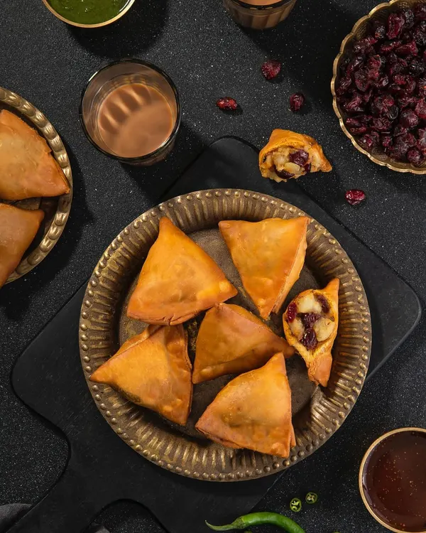 Cranberry-filled samosas, a fusion recipe for US Cranberries India. The dark, moody styling with brass plates and cutting chai glasses gives a rustic yet modern feel.