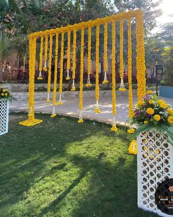 This outdoor Haldi backdrop features a simple frame draped with long strings of yellow marigolds, also known as genda phool. The white latticed pedestals with floral arrangements add a touch of elegance to the sunny, festive setup.