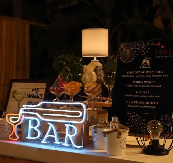 A close-up of one of our chic bar setups. This station features a custom neon sign, a curated drink menu, and stylish decor elements like the table lamp and wire-frame lanterns.