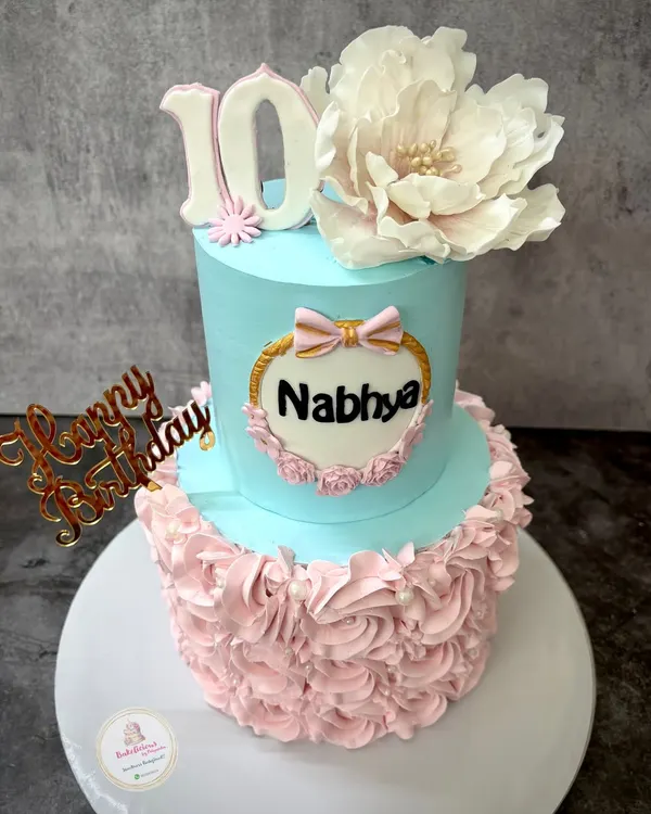 A beautiful two-tier cake for Nabhya's 10th birthday, combining a smooth blue top tier with delicate pink rosettes on the bottom. The large, handmade sugar flower on top adds a touch of elegance.