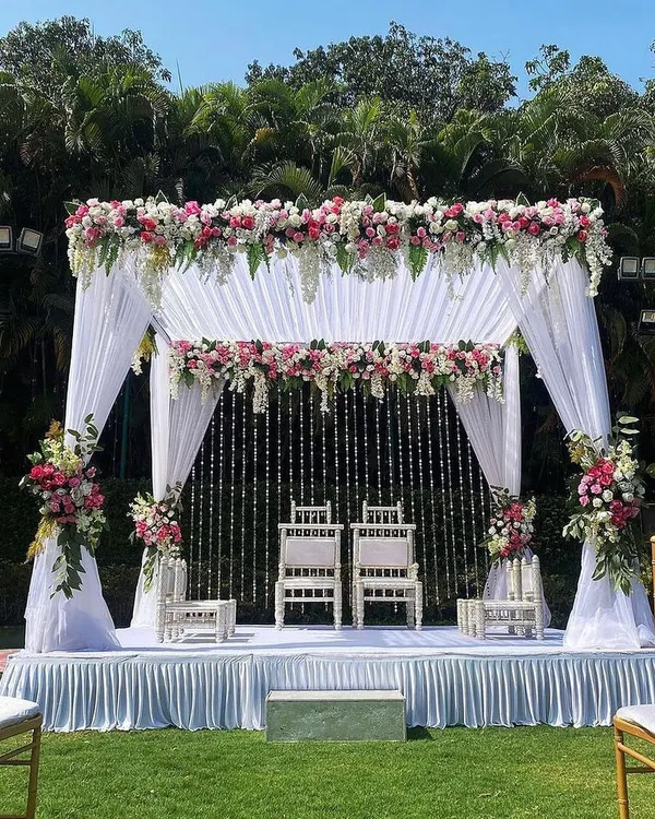 This is an elegant vedi mandap I designed for an outdoor day wedding. The structure is draped in clean white fabric, with lush floral arrangements of pink and white roses on top and along the pillars, creating a modern yet timeless look for the pheras.