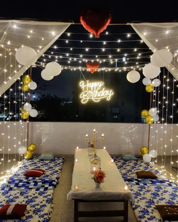 A complete cabana dinner setup on a terrace for a birthday celebration. The structure is draped in sheer fabric and fairy lights, with low seating, candles, and a 'Happy Birthday' neon sign creating a magical dining experience.