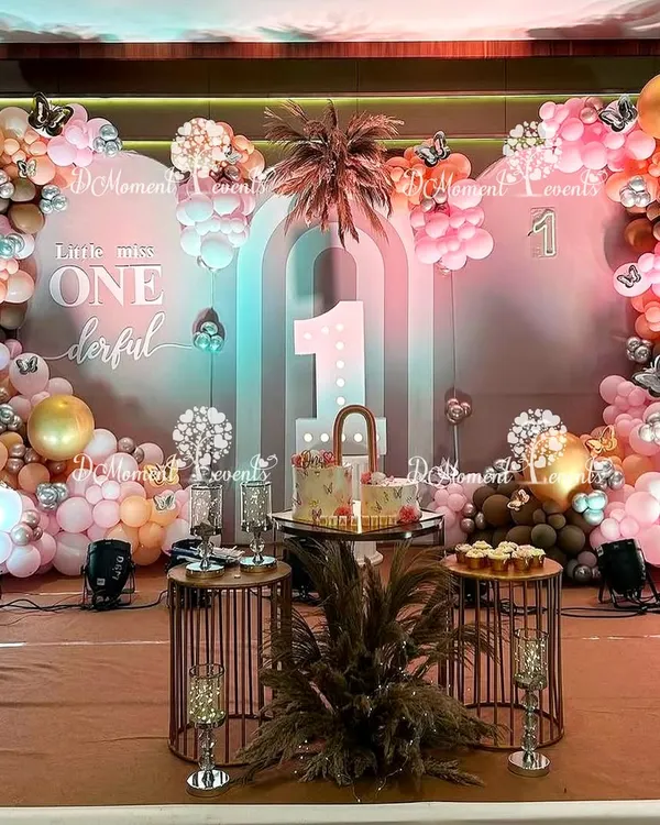 An elegant Boho Rainbow stage for a twins' first birthday, featuring a "Little Miss ONEderful" backdrop, pampas grass, and a sophisticated palette of dusty rose, brown, and gold balloons.