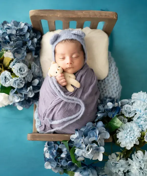 A precious newborn sleeps soundly on a tiny wooden bed, surrounded by a sea of blue flowers. We use creative props and color schemes to design unique newborn portraits.