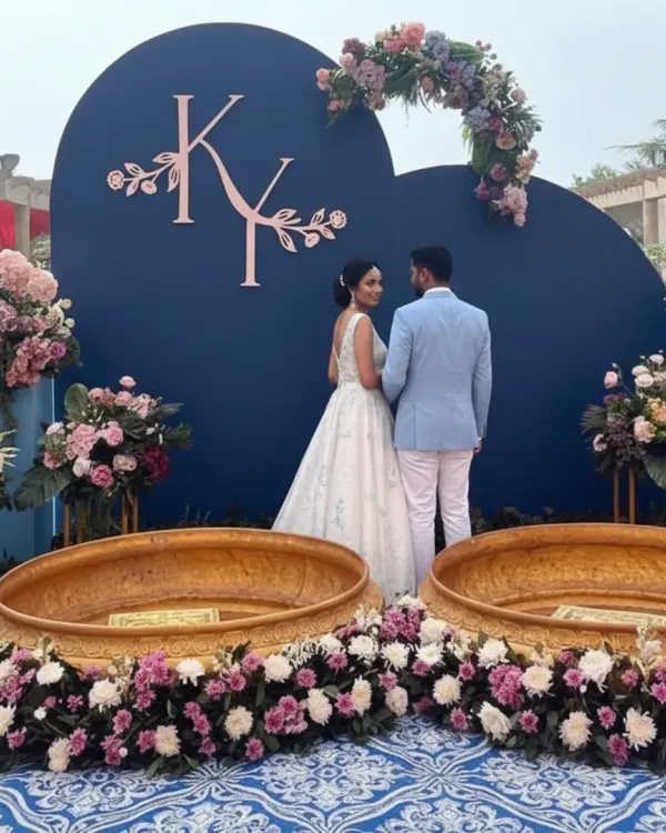 A unique Haldi ceremony setup with a royal blue and pastel theme. The couple is framed by a custom blue backdrop with their initials, surrounded by pink and white floral arrangements and traditional brass urlis.