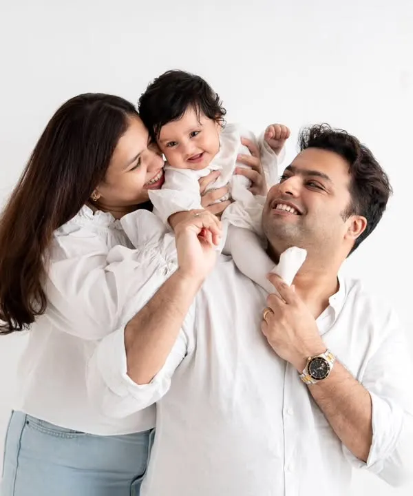 A joyful family of three in a classic studio setting. The baby's giggles are infectious as she sits on her dad's shoulders.