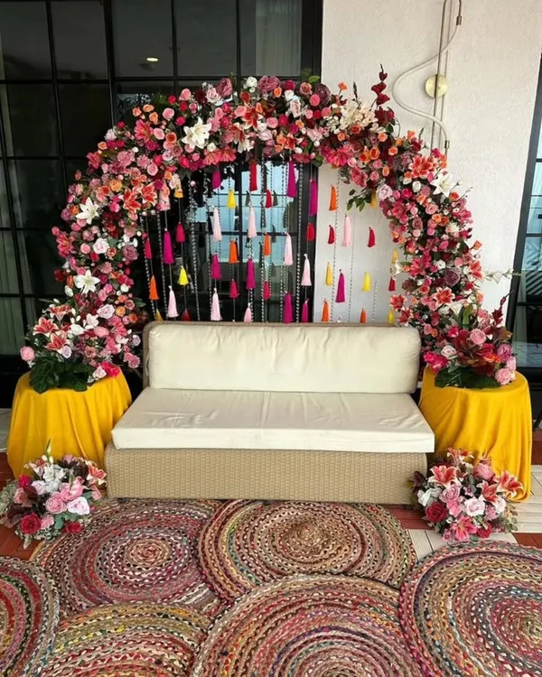 A beautiful seating area I designed for a Mehendi ceremony. The setup is framed by a dense floral arch and a curtain of colorful tassels, with woven rugs adding a bohemian touch.