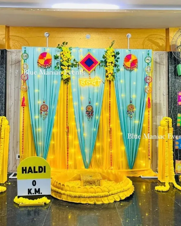 A complete Haldi ceremony setup designed for a home space. The backdrop combines bright yellow and turquoise drapes with traditional hangings and fun, quirky signs like 'Khao Piyo Photo Khichao'. The central seating area is defined by a low, decorated platform, ready for the ceremony.