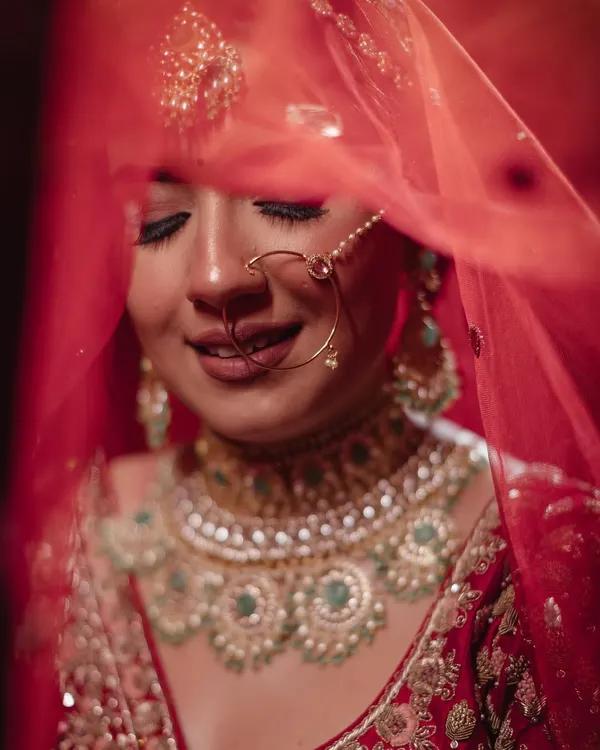 A classic, veiled bridal portrait. The bride's serene smile is captured through her delicate red dupatta, highlighting her stunning jewelry and the joy of the moment.