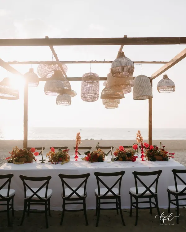 A stunning long-table setup for a beachside dinner in Goa. The scene is set against the ocean, with hanging rattan lamps and vibrant tropical floral runners creating a relaxed yet sophisticated atmosphere.