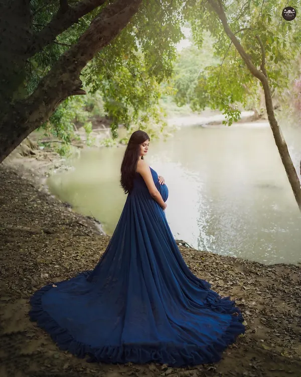 A serene moment captured by the water's edge. The long, flowing train of the blue maternity gown spreads out beautifully, creating a striking contrast with the natural surroundings.