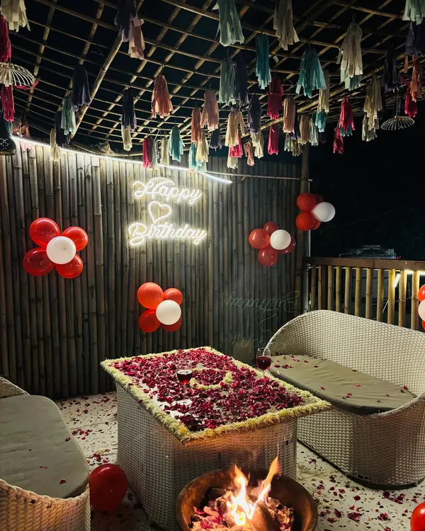 A birthday celebration in full swing. This setup includes a "Happy Birthday" neon sign, balloon decor, and a table covered in rose petals, all centered around a cozy bonfire.