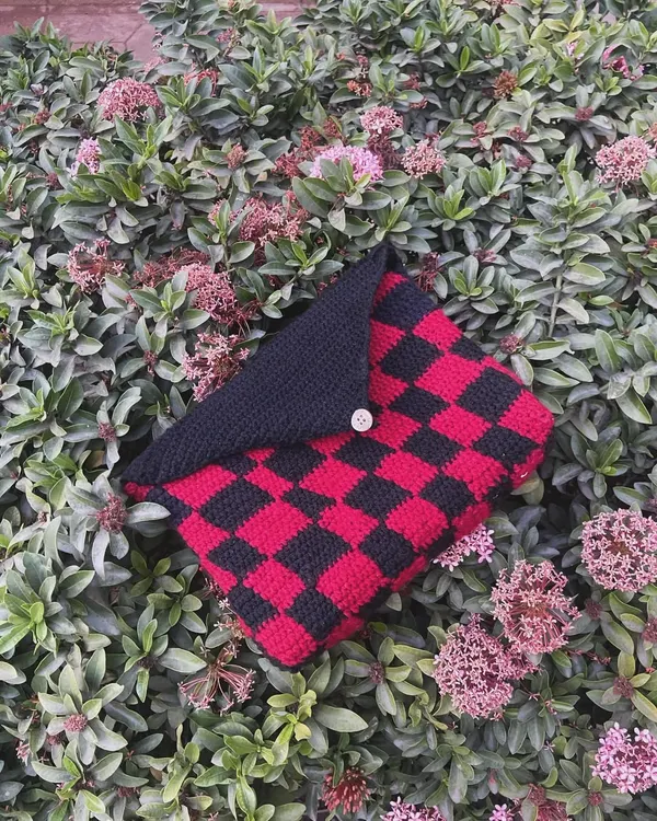 My red and black checkered book sleeve, pictured outside on some bushes. The button flap keeps your book secure inside.