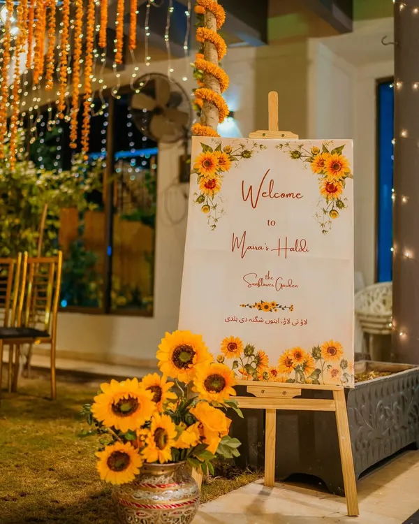 We make sure every detail of your celebration is perfect, day or night. This sunflower-themed welcome sign for an evening Haldi function is complemented by the warm glow of fairy lights and traditional marigold strings.