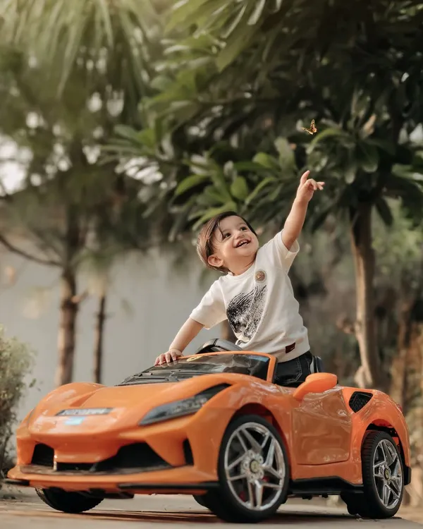 A little driver taking a break from his adventure. This outdoor shot with the orange sports car captures a moment of pure, joyful play against a beautiful green backdrop.