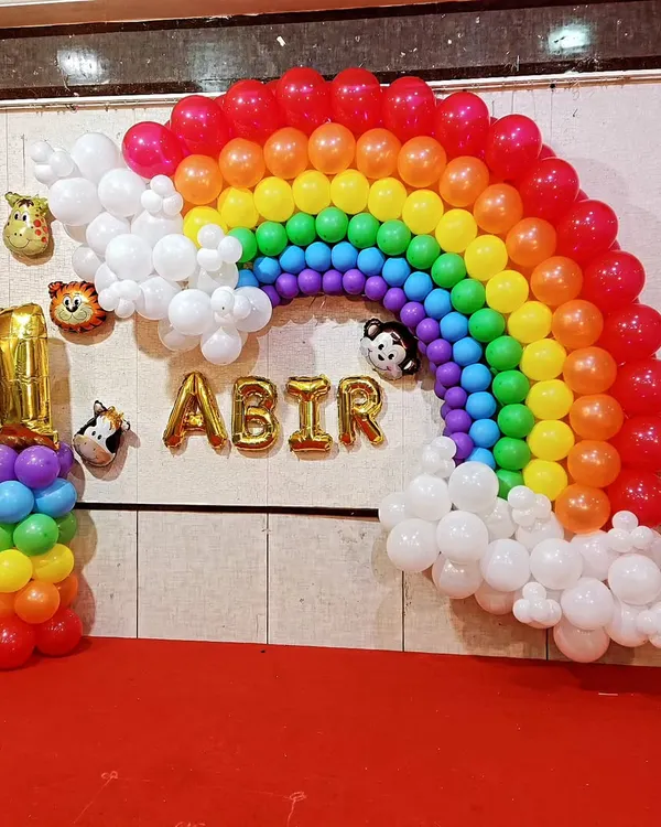 A wall-mounted rainbow balloon arch with fluffy white clouds, creating a beautiful photo backdrop. This setup for a first birthday includes animal head foil balloons peeking out of the clouds and the name "ABIR" in gold letters.