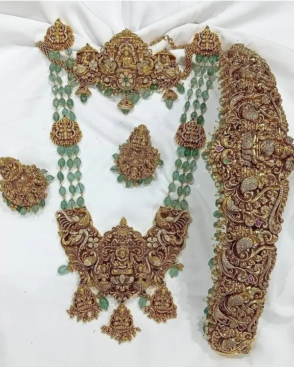 This is a magnificent antique bridal set featuring intricate carvings, goddess motifs, and green bead accents. It includes a choker, a long necklace, a statement vaddanam, and matching earrings.