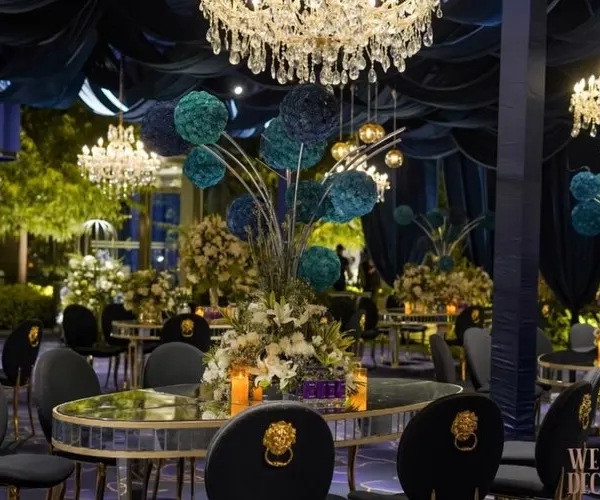 A wide view of the stunning blue and silver themed cocktail setup. The room is bathed in blue light, with dramatic draping, crystal chandeliers, and elegant table settings creating a scene of pure luxury.