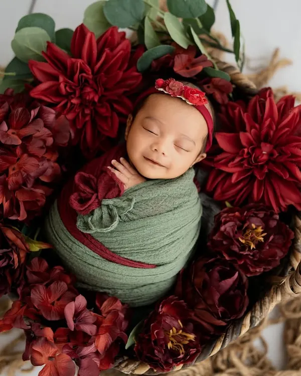 A beautiful smile from this little one, nestled in a basket of rich, red blooms. The contrast between the soft green wrap and the deep red flowers creates a stunning visual.