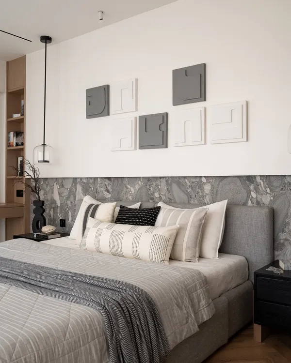 This master bedroom is a serene sanctuary. The bed is framed by an Andromeda leather-finish stone backdrop, complemented by a sleek grey bed and herringbone wood flooring that adds warmth. The textured wallpaper and custom artwork embody quiet luxury in every detail.