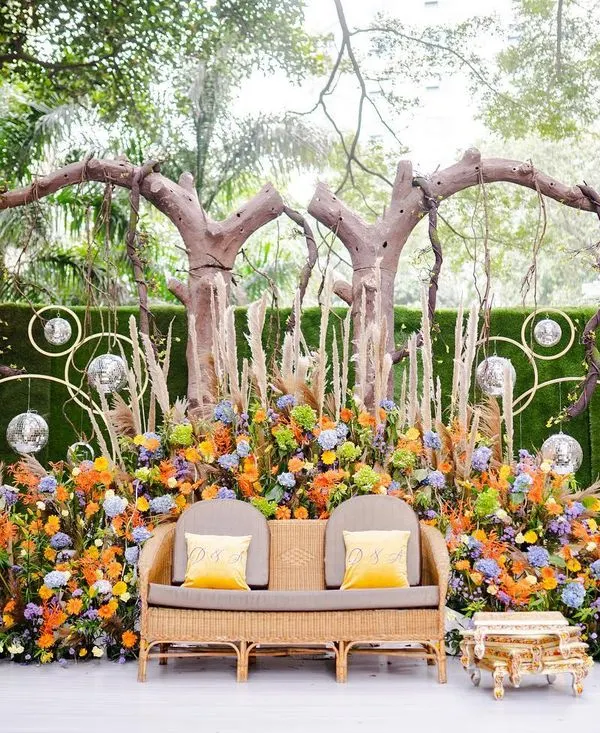 A beautiful and colorful stage for a Mehendi or Haldi ceremony. The design uses a mix of vibrant flowers, pampas grass, and rustic tree branches to create a bohemian and festive look.