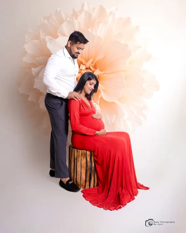 An intimate studio moment between expecting parents. The mom-to-be is seated while her partner stands by her side, set against a simple, elegant giant flower backdrop.