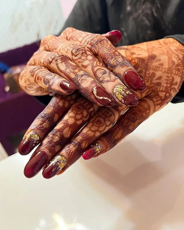 A stunning example of bridal nails designed to enhance the beauty of henna. This set features a rich maroon polish with gold glitter accents that sparkle against the intricate mehndi pattern.
