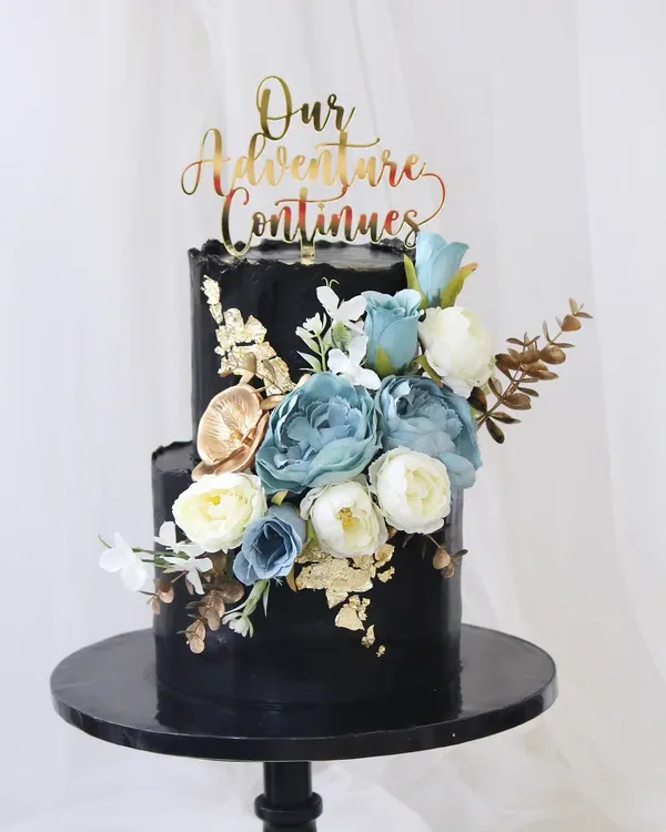A breathtaking black two-tiered wedding cake that exudes modern elegance. The deep black fondant provides a dramatic backdrop for the cascade of blue and white sugar flowers, accented with gold leaf for a touch of luxury.