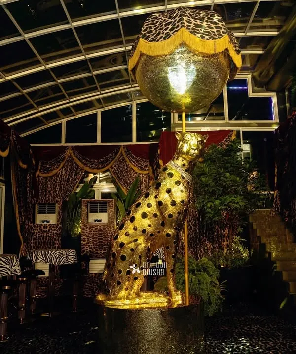 An immersive event space designed with a 'Gilded Instinct' theme, inspired by Roberto Cavalli. The centerpiece is a custom, life-sized golden cheetah statue set against a backdrop of luxurious animal-print drapes and moody lighting, creating an atmosphere of bold opulence.