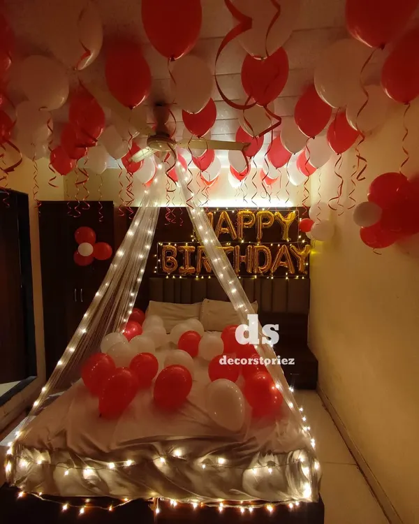 A classic romantic birthday surprise featuring a fairy-light canopy over the bed, which is covered in red and white balloons. The decor is completed with a 'Happy Birthday' foil banner and balloons on the ceiling.