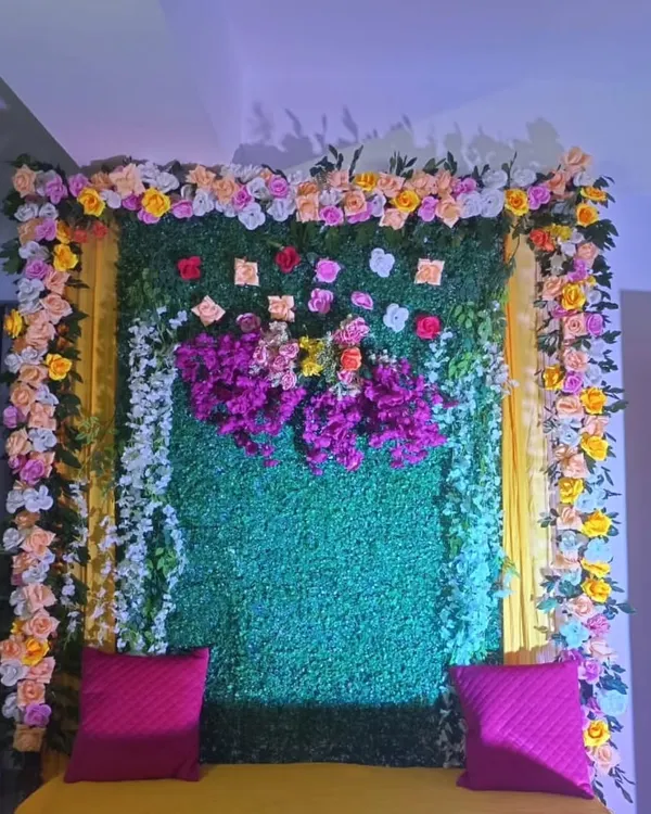 A close-up of a home Haldi backdrop, showing the combination of a green foliage panel with a border of colorful flowers and bright pink cushions.