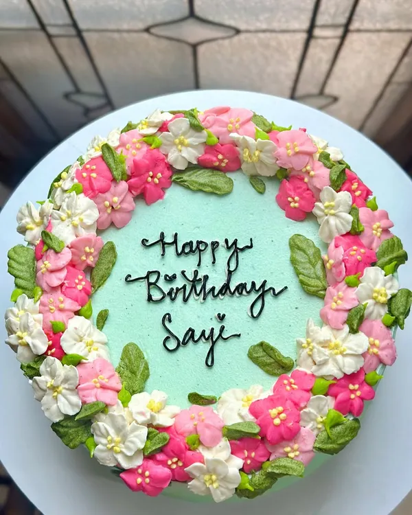 This shows the incredible detail we can achieve with buttercream flowers on a larger cake. This birthday cake for Sayi features a wreath of hand-piped pink and white blossoms on a mint-green base.