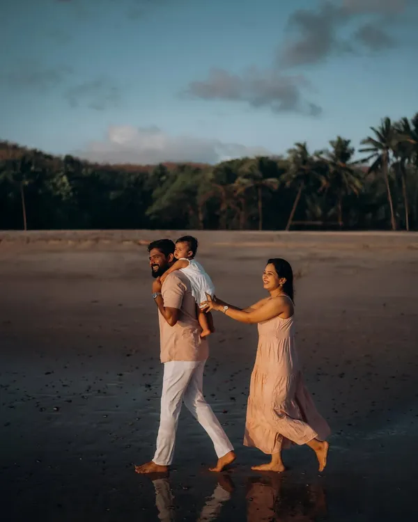 A father gives his child a shoulder ride as they walk along the wet sand at dusk, with the mother holding on playfully. This candid shot captures the simple, joyful moments of a family beach walk.