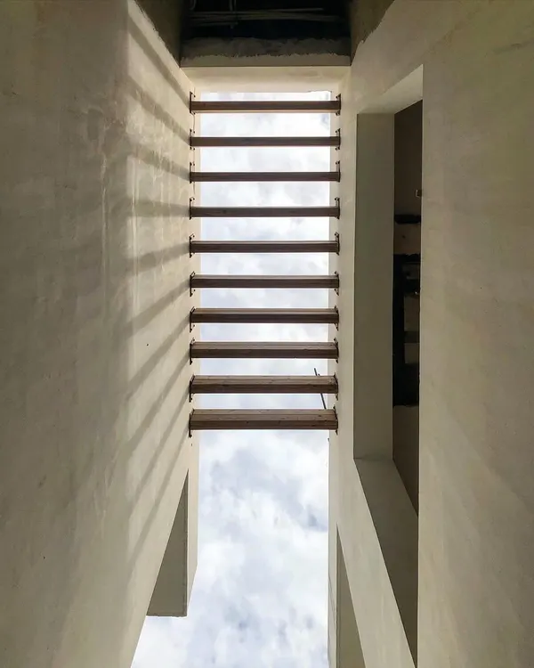 Looking up through a narrow void in a building, where wooden louvers create a rhythmic pattern against the sky. This detail is an example of how we use architectural elements to frame nature and play with light and shadow.