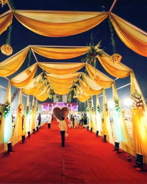 A spectacular wedding entrance walkway with a red carpet, covered by a canopy of golden drapes and lit by spotlights, creating a glamorous path for guests.