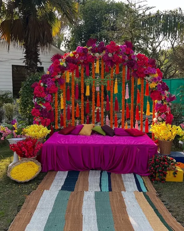 A beautiful outdoor mehndi setup with a vibrant floral arch in shades of pink and red. The decor includes traditional marigold tassels and a comfortable seating area on the lawn.
