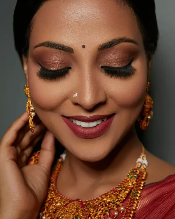 A close-up of a stunning South Indian bridal look. The focus here is on the dramatic, coppery-gold smokey eye and a bold matte lip, which beautifully complements the traditional gold jewelry.