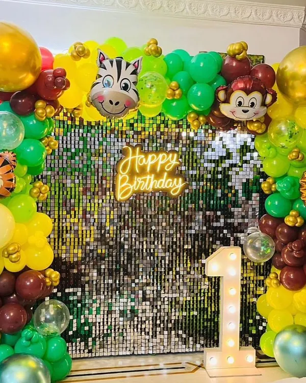 A detailed jungle safari theme for a first birthday, featuring a green and yellow balloon arch with animal foil balloons, a sequin backdrop, and a light-up number "1".